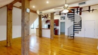Located at 1329 Jefferson St in Lynchburg, VA, this two-story loft unit offers a compelling blend of historic industrial character and contemporary updates. The main floor is generously proportioned, anchored by striking exposed timber posts and beams that speak to the building's original construction. Warm hardwood floors run throughout, complementing the open-concept kitchen and living area. The kitchen is well-appointed with rich cherry cabinetry, granite countertops, stainless steel appliances including an electric range and built-in microwave, and a peninsula with a live-edge wood countertop detail. A dramatic spiral staircase connects the main floor to the upper level, where two loft spaces provide flexible use — one featuring additional exposed timber columns and the other showcasing a whitewashed brick accent wall. The bedroom is tucked under a sloped roofline with exposed beams and a large window that brings in natural light. The bathroom is clean and functional, finished with white tile, a tub-shower combo, and matching cherry vanity. Ceiling fans are installed throughout, and the angled rooflines give each room a distinctive, characterful feel unique to this historic Lynchburg property.