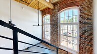 Located at 250 S Depot St in Pendleton, SC, this two-floor loft masterfully blends industrial heritage with contemporary design. The main level opens to a striking living and kitchen area with polished concrete floors, soaring ceilings, and exposed original brick walls. Large arched, multi-pane windows flood the space with natural light while framing views of the surrounding neighborhood. Reclaimed wood ceiling beams add warmth and character throughout. The modern kitchen is outfitted with stainless steel appliances, wood-toned cabinetry, a white quartz island on casters, and a classic subway tile backsplash. A sleek metal staircase with open railings leads to the upper level, where the bedroom and additional living space are situated. Two well-appointed bathrooms serve the home — one featuring warm hardwood floors, a wood-tone vanity with a framed mirror, and a marble-tiled glass-enclosed tub and shower combo, while the other offers polished concrete floors and an elegant marble-surround frameless sliding glass shower. The overall ambiance is a refined industrial aesthetic that honors the building's original character while delivering thoroughly modern comfort and style.