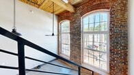 Located at 250 S Depot St in Pendleton, SC, this two-floor loft masterfully blends industrial heritage with contemporary design. The main level opens to a striking living and kitchen area with polished concrete floors, soaring ceilings, and exposed original brick walls. Large arched, multi-pane windows flood the space with natural light while framing views of the surrounding neighborhood. Reclaimed wood ceiling beams add warmth and character throughout. The modern kitchen is outfitted with stainless steel appliances, wood-toned cabinetry, a white quartz island on casters, and a classic subway tile backsplash. A sleek metal staircase with open railings leads to the upper level, where the bedroom and additional living space are situated. Two well-appointed bathrooms serve the home — one featuring warm hardwood floors, a wood-tone vanity with a framed mirror, and a marble-tiled glass-enclosed tub and shower combo, while the other offers polished concrete floors and an elegant marble-surround frameless sliding glass shower. The overall ambiance is a refined industrial aesthetic that honors the building's original character while delivering thoroughly modern comfort and style.