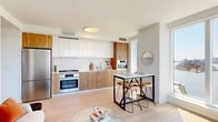 Located at 20 Hallets Point in Astoria, Queens, this modern residence offers four bedrooms and one bathroom across a thoughtfully designed single-floor layout. Light hardwood flooring runs throughout, tying together the clean, contemporary aesthetic that defines the space.

The open-concept kitchen is a standout feature, combining high-gloss white upper cabinetry with warm wood-grain lower cabinets and a sleek white stone countertop and backsplash. Stainless steel Bosch appliances, including a gas range, oven, and refrigerator, complete this well-appointed culinary space. A bar-height dining table adjacent to the kitchen provides a casual dining option.

The living room is bathed in natural light through floor-to-ceiling windows that frame sweeping views of the East River and the Manhattan skyline — a truly dramatic focal point of the home. Light-toned walls and pale hardwood floors amplify the brightness throughout.

The welcoming hallway features recessed lighting and built-in storage, with sightlines that draw the eye toward the city views beyond. The primary bedroom is comfortably proportioned with a large closet and serene, neutral finishes. The bathroom is finished in a clean, modern style with warm wood vanity cabinetry, a backlit mirror, white tile surround, and a soaking tub.