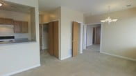 This single-floor residential unit offers a straightforward and functional layout designed to maximize usability. The interior is finished with neutral beige carpeting and warm-toned walls, creating a clean and cohesive aesthetic throughout. The central hallway serves as the spine of the unit, connecting multiple rooms and providing excellent flow between spaces. Notable storage is a key feature, with generously sized bi-fold closet doors offering ample wardrobe or linen storage space. Additional doors lead to what appear to be bedrooms, each accessible directly from the hallway, ensuring privacy and separation between living areas. A partial view of the kitchen area is visible, featuring a countertop pass-through that connects it to the adjacent living space, adding a sense of openness. The unit benefits from recessed and overhead lighting fixtures that illuminate each area effectively. Floor vents throughout suggest a forced-air heating and cooling system for year-round comfort. White baseboards and door trim provide clean contrast against the carpeted floors. The overall ambiance is practical and move-in ready, offering a well-organized residential layout suited for everyday living.