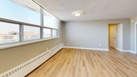 Located on the 16th floor at 25 Leith Hill Road in Toronto's North York neighbourhood, this unit offers a thoughtfully designed open-concept layout that combines the kitchen and living area into one cohesive space. The kitchen is a standout feature, outfitted with sleek white cabinetry, clean white countertops, a white tile backsplash, and stainless steel appliances including a refrigerator, electric range, and dishwasher. A white kitchen island provides additional prep and counter space, while light-toned tile flooring ties the kitchen area together with a bright, clean aesthetic. The living area transitions seamlessly from the kitchen and is finished with warm light hardwood flooring that adds a natural contrast to the neutral grey walls. Large floor-to-ceiling windows line the exterior wall, flooding the space with abundant natural light and offering impressive panoramic views of the Toronto skyline and surrounding residential neighbourhoods. A baseboard heating unit runs along the window wall, ensuring year-round comfort. A private balcony door provides direct outdoor access, further enhancing the connection to the city views. The textured ceiling and flush-mount light fixtures complete the practical yet polished interior. This unit presents a clean, modern living environment well-suited for urban living.