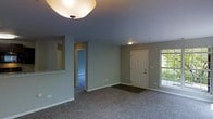 This single-floor residential space offers a clean, functional layout designed for comfortable everyday living. The main living area is open and spacious, featuring wall-to-wall carpeting in a neutral tone that adds warmth underfoot. Soft gray walls provide a versatile backdrop throughout, complementing the white trim and baseboards that frame each room neatly.

Ceiling-mounted light fixtures provide consistent ambient lighting, while a side window in the living area allows natural light to filter in, brightening the space. The front entry is clearly defined, featuring a classic paneled white door with a dark entry mat, creating a welcoming transition from outdoors to indoors.

A pass-through opening on one side hints at a connected kitchen area, where light-toned countertops are visible, suggesting a practical and efficiently designed culinary space. A hallway leading off the main room provides access to additional rooms beyond, contributing to the overall sense of a well-organized floor plan.

The overall ambiance of the space is understated and move-in ready, with a neutral color palette and consistent finishes throughout that offer a solid foundation for personalization. The single-floor layout ensures ease of navigation and accessibility across all areas of the home.