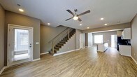 This two-story space offers a well-designed open-concept main level that seamlessly connects the living area to the kitchen. The living room is spacious and bright, finished with wood-look plank flooring that runs throughout the main level, providing a warm yet contemporary feel. Neutral gray walls create a versatile backdrop, complemented by white baseboards that add a clean, finished look.

Recessed lighting is distributed across the ceiling, ensuring consistent illumination throughout the open area, while a ceiling fan with an integrated light fixture adds both function and a decorative element. A staircase with modern metal balusters and wood-toned handrails leads to the upper level, serving as a subtle architectural focal point within the living space.

Toward the rear, a glimpse of the kitchen reveals a countertop bar area and pendant lighting, hinting at a well-appointed cooking and dining space. A window at the back of the home allows natural light to filter into the area. The overall ambiance is clean, modern, and move-in ready, with cohesive finishes that carry throughout the visible areas of the main floor.