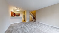 This two-story residence offers a thoughtfully designed layout spread across two levels. The main floor centers around an open-concept living and kitchen area, where plush wall-to-wall carpeting defines the generous living space. The kitchen is well-equipped with rich dark wood cabinetry, granite countertops, stainless steel appliances including a refrigerator, dishwasher, and range, all set atop light hardwood flooring. A breakfast bar provides a seamless connection between the kitchen and living area. A staircase with white railings leads to the upper level, maintaining an open and airy feel throughout. The bathrooms are finished with matching granite-topped vanities, warm wood cabinetry, and clean tile flooring. A full bathroom on the upper level includes a walk-in glass-door shower. The bedrooms are carpeted and bathed in natural light through large windows with views of the surrounding neighborhood. One bedroom offers direct access to a private balcony through sliding glass doors, providing an outdoor retreat. Neutral warm beige tones are carried consistently throughout the space, creating a cohesive and inviting ambiance well-suited for comfortable everyday living.