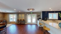 Located at 231 Calibre Chase Dr in Raleigh, NC, this single-floor community clubhouse offers a thoughtfully designed gathering space that blends functionality with inviting style. Rich hardwood flooring flows throughout, creating a warm and cohesive foundation across all areas of the space.

The open layout connects a comfortable lounge area to a well-appointed kitchen and coffee station. The lounge features leather armchairs arranged around a round coffee table atop a woven rug, curated artwork including a striking reclaimed wood geometric wall piece, and a dedicated workspace with a desk and computer — making it equally suited for relaxation or productivity. Neutral gray walls and recessed lighting contribute to a calm, modern ambiance.

The kitchen area is a standout, showcasing bold navy blue subway tile that creates a dramatic focal point against crisp white cabinetry and light quartz countertops. Stainless steel appliances including a full-size refrigerator and built-in microwave add practicality, while bar-height stools along the peninsula invite casual interaction.

Adjacent to the kitchen, a dedicated coffee bar with a built-in espresso machine invites residents to gather and connect. French doors along one wall flood the space with natural light and open to views of an outdoor pool area, seamlessly extending the welcoming atmosphere beyond the interior.