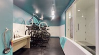 This purpose-built bike repair and storage room is designed with cyclists in mind, offering a well-organized and functional space for both storing and maintaining bicycles. The room features a striking two-tone color scheme, with crisp white lower walls contrasted by bold teal upper walls and a matching teal ceiling, creating a vibrant and energetic atmosphere. Recessed lighting illuminates the space evenly, ensuring excellent visibility for repair work at any time of day. A multi-level black metal bike rack system serves as the centerpiece, capable of accommodating multiple bicycles in a compact and efficient arrangement. A dedicated work surface along one wall provides a convenient area for performing maintenance and repairs. The dark rubberized flooring is both durable and easy to clean, making it well-suited for the demands of a high-use utility space. A large glass partition or window on one side allows for natural light to filter in while maintaining visual connectivity with the adjacent area. The room is thoughtfully appointed to meet the practical needs of avid cyclists, combining smart storage solutions with a workspace that makes routine bike maintenance straightforward and accessible.