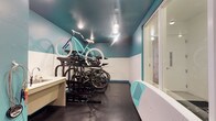 This purpose-built bike repair and storage room is designed with cyclists in mind, offering a well-organized and functional space for both storing and maintaining bicycles. The room features a striking two-tone color scheme, with crisp white lower walls contrasted by bold teal upper walls and a matching teal ceiling, creating a vibrant and energetic atmosphere. Recessed lighting illuminates the space evenly, ensuring excellent visibility for repair work at any time of day. A multi-level black metal bike rack system serves as the centerpiece, capable of accommodating multiple bicycles in a compact and efficient arrangement. A dedicated work surface along one wall provides a convenient area for performing maintenance and repairs. The dark rubberized flooring is both durable and easy to clean, making it well-suited for the demands of a high-use utility space. A large glass partition or window on one side allows for natural light to filter in while maintaining visual connectivity with the adjacent area. The room is thoughtfully appointed to meet the practical needs of avid cyclists, combining smart storage solutions with a workspace that makes routine bike maintenance straightforward and accessible.