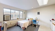 This studio apartment on the fifth floor of 206 Dryden Road in Ithaca, New York, offers a compact and functional open-plan layout designed to maximize every square foot. The main living area serves as both a sleeping and living space, featuring a large picture window that frames views of the surrounding Ithaca cityscape and fills the room with natural light. A skylight overhead further brightens the interior, complementing the clean white walls and light-toned ceiling. Carpeted flooring in the living and sleeping zone adds warmth underfoot, while the entry and kitchen area transition to a smooth hard-surface floor.

The kitchenette is neatly integrated along one wall, equipped with white cabinetry featuring wood-trimmed accents, ample upper and lower storage, a full-size refrigerator, a range with stovetop, a sink, and space for a microwave. A dedicated study area with a built-in desk and chair provides a practical workspace. Additional storage is accommodated by a tall bookcase and a multi-drawer dresser.

The private bathroom includes a single-sink vanity with a wood-tone cabinet, a wide wall mirror, and a shower-tub combination with sliding glass doors. The unit's efficient design and elevated position make it a well-appointed option in the heart of Ithaca.