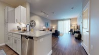 This well-appointed single-floor residence offers a thoughtfully designed open-concept layout that seamlessly connects the kitchen, dining, and living areas. The kitchen features crisp white cabinetry, a full suite of stainless steel appliances including a refrigerator, range, and microwave, and a generously sized granite-topped island that doubles as a breakfast bar. Hardwood-style flooring runs throughout the main living areas, adding warmth and visual continuity.

The living room is bright and inviting, anchored by large floor-to-ceiling sliding glass doors dressed with sheer vertical blinds that allow abundant natural light to fill the space. Track lighting overhead provides additional illumination, complementing the room's airy, contemporary ambiance.

The bedroom is a calm retreat finished with plush wall-to-wall carpeting, neutral toned walls, and windows fitted with blinds and sheer curtains that invite soft, diffused light. The room offers generous proportions suitable for a full bedroom suite.

The bathroom features wood-look vinyl flooring, clean white paneled doors, and a tub, offering a functional and tidy finish. A dedicated entryway near the kitchen provides a practical transition point into the home. Throughout, neutral grey and beige tones create a cohesive, modern aesthetic.