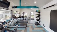 Located at 1231 W Francisquito Ave in West Covina, California, this single-floor fitness center is thoughtfully designed to support a well-rounded workout experience. The space features vaulted ceilings accented by bold teal structural beams, lending a modern and energetic aesthetic to the room. Recessed lighting and large windows along multiple walls flood the interior with natural light and offer views of the surrounding outdoor area, including a pool visible from the weight training zone.

The fitness center is equipped with a comprehensive selection of cardio machines, including multiple treadmills and elliptical trainers by Matrix, as well as a spin bike. A dedicated free weights area includes a full rack of dumbbells, a weight bench, and a selection of kettlebells and medicine balls stored on wall-mounted racks. A wall-mounted flat-screen television adds entertainment for those engaged in longer cardio sessions.

The flooring features a geometric patterned carpet in shades of teal, gray, and blue, complementing the room's color palette. A glass door provides direct access to the outdoor grounds. A wall-mounted sanitizer dispenser and a split-system air conditioning unit are also present, ensuring a clean and comfortable environment for all users.