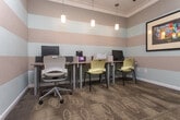 Located at 500 McDaniel St SW in Atlanta, Georgia, this business center offers a well-appointed, single-floor workspace designed for productivity and comfort. The space is immediately distinguished by its bold horizontal striped walls, alternating between warm taupe and soft sage tones, creating a visually dynamic yet calming environment.

The interior is furnished with multiple workstations equipped with desktop computers, keyboards, and a shared printer, making it a fully functional workspace for everyday computing needs. Pendant lighting fixtures hang from the ceiling, casting a warm, focused glow over the desks, while crown molding adds a refined architectural detail to the room.

Patterned carpet flooring with a subtle circular motif grounds the space, complementing the color palette of the walls. Dark-framed glass doors with sidelights provide both access and a sense of transparency between areas, while framed artwork — including a notable license plate map of the United States — adds personality and a touch of Americana to the walls.

The layout accommodates multiple users simultaneously, with ergonomic seating and ample desk surface area. Safety features, including a fire alarm and security camera, are also present. The overall ambiance is professional yet approachable, offering a comfortable and well-equipped environment for residents or visitors needing a dedicated workspace.