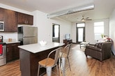This single-floor unit is designed with a contemporary yet warm aesthetic, combining functional layouts with cohesive finishes across every room. Rich hardwood-style flooring runs throughout the space, providing a consistent foundation that ties each area together seamlessly.

The living room is a welcoming, light-filled space anchored by large windows that allow natural light to pour in. A ceiling fan overhead adds comfort, while the open layout accommodates generous seating and a rustic industrial coffee table on casters.

The kitchen is a standout feature, offering dark espresso cabinetry paired with light gray countertops and a full suite of stainless steel appliances, including a range, microwave, refrigerator, and dishwasher. Open metal shelving adds a modern industrial touch, and a central island provides additional prep and seating space.

The bedroom is comfortably sized with multiple windows dressed in blinds and curtains for privacy and light control. A ceiling fan, hardwood flooring, and a sliding barn-style closet door contribute to the room's relaxed, functional character.

The bathroom connects directly to the bedroom via a hallway, featuring a clean white vanity and straightforward, modern fixtures. The overall ambiance of the unit is calm and cohesive, with neutral tones and warm wood elements creating an inviting, livable environment.
