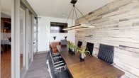 This single-floor professional space is designed with a clean, contemporary aesthetic that balances functionality with refined style. The centerpiece of the space is a well-appointed conference room that immediately commands attention with its distinctive reclaimed wood-plank accent wall, which adds warmth and texture against the cool gray tones of the surrounding walls. A large, dark-stained wooden conference table anchors the room, surrounded by quilted black leather executive chairs that convey a polished, professional atmosphere. Overhead, a sleek rectangular pendant light fixture provides focused illumination, enhancing the room's modern character. Large windows along one wall flood the space with natural light and offer views of the surrounding streetscape, creating an open and connected feel. A wall-mounted flat-screen display is thoughtfully positioned for presentations and collaborative meetings. Beyond the conference room, glass-paneled sliding doors open to additional areas, including what appears to be a kitchenette and further office or lounge spaces, suggesting a well-organized and fully equipped professional environment. The overall ambiance is sophisticated and inviting, making it well-suited for productive meetings and client engagements.