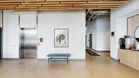 Located at 1624 W Babcock St in Bozeman, Montana, this contemporary lobby serves as the welcoming hub of the Haymaker Apartments. The space is defined by its striking architectural design, where a distinctive slatted wood ceiling adds warmth and texture, creating a refined industrial aesthetic that is complemented by exposed ductwork and polished concrete floors throughout.

Floor-to-ceiling glass curtain walls line the street-facing façade, flooding the interior with abundant natural light and offering open views of the surrounding Bozeman streetscape. The main lounge area features a comfortable seating arrangement with modern upholstered armchairs gathered around a round coffee table, providing residents and guests with an inviting place to gather.

A dedicated coffee station, outfitted with a coffee maker and light wood cabinetry, is accented by a large round mirror and offers a convenient amenity near the building entrance. Adjacent casual dining seating with contemporary chairs and a round table adds further functionality to the space.

A wide corridor leads to an elevator and access doors with deep green painted finishes, contributing to the cohesive modern color palette. Carefully curated wall art enhances the aesthetic throughout. The overall ambiance is one of clean, contemporary design that balances industrial elements with natural warmth, creating an upscale and functional first impression for residents and visitors alike.