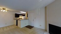 This single-floor residence offers a practical and comfortable layout designed to maximize everyday functionality. The open-concept design connects the living area to the kitchen, creating a cohesive and welcoming atmosphere throughout the space. The carpeted living area provides a soft, warm underfoot feel and flows naturally toward the main entry door, which is framed by neutral-toned walls that lend a clean and understated aesthetic. The kitchen is a standout feature, showcasing dark espresso-finished cabinetry that creates a bold contrast against the lighter walls and countertops. Equipped with essential appliances including a refrigerator, range with overhead ventilation, and ample counter space, the kitchen is both stylish and highly functional. A peninsula-style counter serves as a natural divider between the kitchen and the living area, offering additional workspace as well as a casual dining or gathering spot. Overhead lighting in the kitchen ensures the space is well-lit and practical for meal preparation. The entry area is accented by a dark floor mat, providing a defined transition point into the home. Overall, the space presents a tidy, move-in-ready feel with a cohesive color palette and efficient use of its layout.