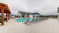 Located at 200 Villas Dr in Lavon, Texas, this generously sized community amenity center spans a single level and offers a resort-inspired outdoor experience. At the heart of the facility is a large, sparkling swimming pool with clearly marked depth designations and a dedicated shallow wading area lined with in-pool lounge chairs — ideal for relaxing in the water. The expansive stone-tiled sundeck surrounds the pool on all sides, providing ample space for rows of modern sling-style chaise lounges and umbrella-shaded seating areas. A covered pavilion with a dark-stained wood ceiling, ceiling fans, and open-air columns offers a shaded retreat directly adjacent to the pool deck, while a separate wood pergola on the opposite end provides additional outdoor gathering space. The main amenity building is clad in crisp white horizontal siding with a dark pitched roof, housing restroom facilities, and a fitness or leasing studio accessible from the pool deck. Exterior amenities include a drinking fountain station, wall-mounted lighting, and gated perimeter fencing that encloses the entire complex. The surrounding neighborhood views and open sky contribute to the bright, airy atmosphere of this well-appointed community recreation destination.