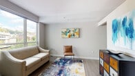 This single-floor unit presents a well-organized layout that balances modern design with practical functionality throughout its spaces. The living area is bright and welcoming, benefiting from a large window that frames outdoor greenery and fills the room with natural light. Light-toned hardwood-style flooring, neutral gray walls, and colorful abstract artwork contribute to a contemporary yet relaxed atmosphere, complemented by a vibrant multi-colored area rug.

The kitchen is a standout feature, outfitted with rich dark-stained cabinetry that extends to the ceiling for generous storage. Stainless steel appliances, including a refrigerator, microwave, and range, pair with a white subway tile backsplash and dark countertops for a sleek, modern aesthetic. A built-in breakfast bar with modern barstools provides a casual dining option.

The bedroom maintains the unit's calm, neutral palette with soft carpeting, subtle tray ceiling detailing, and coordinating abstract wall art, creating a restful retreat. The bathroom features large-format tile flooring, white cabinetry with clean chrome hardware, an integrated sink, a wide mirror, and a tiled shower enclosure, delivering a straightforward and functional design throughout.