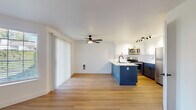 This single-floor residence offers a thoughtfully designed open-concept layout finished with light hardwood flooring throughout. The living area serves as the heart of the space, anchored by a tile-surround fireplace and bathed in natural light from a large window and a sliding glass door that opens to an outdoor area. A ceiling fan with an integrated light fixture provides comfort and airflow, while vertical blinds offer adjustable privacy.

The kitchen is a standout feature, distinguished by a bold navy-blue island with a white quartz countertop and an undermount sink fitted with a matte black faucet. Upper cabinetry is finished in crisp white with black hardware, while lower cabinets mirror the navy tones of the island. A geometric hexagon-tile backsplash adds texture, and stainless steel appliances — including a range, over-the-range microwave, and refrigerator — complete the space. Track lighting and an adjacent open pantry with built-in shelving enhance functionality.

The bathroom features a gray shaker-style vanity, white countertop, round mirror with a black frame, and a tub-shower combination. A bedroom with a baseboard heater, natural light from a window, and access to a closet rounds out the layout.