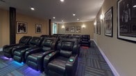 Located at 300 Canal in Lawrence, Massachusetts, this single-floor screening room delivers an immersive, theater-style experience within a well-appointed interior setting. The room is designed to accommodate a sizeable audience, with multiple rows of plush black leather recliner seats arranged in a tiered layout to ensure unobstructed sightlines from every position. Each seat features built-in LED accent lighting in blue and teal hues, lending the space a cinematic atmosphere reminiscent of a commercial movie theater.

The warm beige walls are adorned with a curated collection of framed classic movie posters, adding a sophisticated, film-enthusiast aesthetic throughout. Recessed ceiling lighting and wall-mounted sconces provide layered illumination that can set the mood for any screening. The flooring consists of patterned carpet tiles in complementary tones of gray, blue, and teal, contributing both visual interest and acoustic dampening.

At the front of the room, a large wall-mounted flat-screen display serves as the primary viewing screen, supported by a rustic wooden media console. Dark structural columns punctuate the space, and a clearly marked exit door provides access to an adjacent corridor. The overall ambiance is polished, comfortable, and purpose-built for an elevated viewing experience.