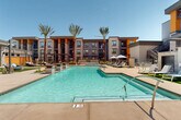 Located at 575 S Cotton Ln in Goodyear, Arizona, this outdoor amenity space showcases a resort-style pool courtyard designed to deliver a premium leisure experience in the heart of the Sonoran Desert. The centerpiece is a generously sized swimming pool with crystal-clear blue water, framed by a broad, sun-drenched concrete deck ideal for relaxation and socializing. Rows of cushioned lounge chairs and market umbrellas are thoughtfully arranged around the pool's perimeter, providing ample shaded and open-air seating options. Towering palm trees line the courtyard, adding a tropical ambiance that complements the warm Arizona climate. A spa or water feature is also present, offering a more intimate retreat alongside the main pool. The surrounding multi-story building features a contemporary architectural design, blending stucco, stone cladding, and bold accent colors including orange and deep blue, creating a vibrant and modern aesthetic. A pergola-shaded outdoor lounge area with additional seating and a grilling station is tucked along one side of the courtyard, perfect for gatherings. The space is enclosed by the building on multiple sides, creating a semi-private, resort-like atmosphere bathed in natural sunlight under open Arizona skies.
