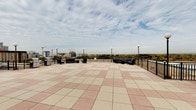Located at 4400 East-West Hwy in Bethesda, Maryland, this rooftop-level space offers a compelling combination of open-air amenities and polished interior transitional areas. The expansive rooftop terrace is finished with a geometric pattern of light and rose-toned pavers, creating a spacious and well-appointed outdoor environment. Anchored by globe-style lampposts, the terrace is equipped with multiple seating arrangements featuring dark-toned outdoor furniture, as well as dedicated grilling stations ideal for gatherings. A perimeter safety railing frames sweeping panoramic views of the Bethesda skyline, with surrounding urban development and lush tree canopies visible on the horizon beneath open skies. Transitioning indoors, the elevator lobby is distinguished by floor-to-ceiling marble-tiled wall cladding in soft grey tones, complemented by blue-grey carpeting and warm wall sconce lighting. Large windows in the vestibule area maintain a visual connection to the outdoor terrace, allowing natural light to filter into the interior corridor. Access to the pool and additional building amenities is provided via clearly marked doorways within the carpeted hallway. Together, the rooftop terrace and its adjoining interior spaces deliver a refined, amenity-rich experience well-suited to a prominent Bethesda address.
