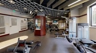 Located at 4405 East-West Highway in Bethesda, MD, this fitness center offers a comprehensive single-floor workout facility designed with both functionality and aesthetics in mind. The space features a distinctive ceiling design with an open grid framework accented by recessed and linear lighting, creating a well-lit environment throughout. Rich mahogany-toned columns and wall accents contrast with neutral walls, lending the space a polished, upscale character.

The cardio zone accommodates a generous row of treadmills, stationary bikes, ellipticals, and stair climbers, with mounted flat-screen TVs positioned for easy viewing during workouts. Full-length mirrors line multiple walls, enhancing the sense of openness and allowing users to monitor their form. The free weight area includes a well-organized rack of dumbbells, barbells, and benches, complemented by built-in shelving stocked with yoga mats and exercise accessories. A dedicated strength training section features a variety of resistance machines. The rubber-floored surfaces throughout provide safety and durability. Natural light filters in through exterior-facing windows and glass doors, connecting the interior to the outside. The overall ambiance is professional, clean, and purposefully arranged to support a full range of fitness activities.