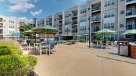 Located at 91 York Rd in Willow Grove, PA, this outdoor amenity space serves as a central gathering hub set within a multi-story residential complex. The centerpiece is a generously sized swimming pool with crystal-clear turquoise water, enclosed by a decorative wrought iron fence for safety and security. A row of blue lounge chairs lines the pool deck, offering ample space for sunbathing and relaxation.

Adjacent to the pool, a wide concrete courtyard provides multiple seating areas outfitted with white tables and chairs shaded by striped green-and-white umbrellas. String lights are strung between poles above the courtyard, creating an inviting atmosphere for evening gatherings. A brick-accented outdoor grilling station with a stainless steel grill serves as a social focal point, complemented by a ping-pong table nearby for recreation.

A charming wooden pergola with lattice detailing anchors one corner of the space, providing a shaded retreat alongside a lush green turf lawn area with additional lounge seating. Manicured landscaping with shrubs and plantings borders the space throughout. The entire area is framed by the surrounding multi-story building, whose balconies overlook the courtyard, reinforcing the sense of a vibrant, well-appointed community outdoor living space.