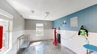 Located at 86–94 Mason Dr in Red Bank, TN, this single-room amenity space is thoughtfully designed to serve two distinct functions: a pet spa and a bike repair station. The room features a polished concrete floor that is both durable and easy to clean, making it well-suited for its active, utility-focused purposes. White subway tile lines the lower half of the walls, transitioning to a two-tone color scheme of teal and gray on the upper portion, lending the space a clean, contemporary aesthetic.

The pet spa area is equipped with a stainless steel grooming tub, complete with steps for easy pet access, integrated plumbing fixtures, and a wall-mounted pet dryer. A stainless steel utility table provides additional workspace, and posted signage outlines pet spa rules for user guidance. The bike repair station features a prominent red floor-standing bike repair stand alongside mounted tools, offering residents a fully functional maintenance area. A wall-mounted air pump and posted bike repair signage complete this section.

Natural light enters through a window framed in white, brightening the interior alongside flush-mounted fluorescent ceiling fixtures. A bold red-framed glass door serves as the primary entry point, adding a striking design accent. The space is well-ventilated with visible HVAC registers throughout.