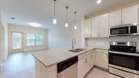 This single-story residence at 659 Treeland Dr in Ladson, South Carolina presents a clean, modern interior with a thoughtfully designed open-concept layout. The kitchen serves as the centerpiece of the home, anchored by a large island with granite countertops, an undermount sink, and a sleek pull-down faucet. Pendant lighting hangs above the island, complementing the recessed ceiling lights throughout. Crisp white cabinetry extends to the upper walls, providing generous storage, while a full suite of stainless steel appliances—including a French door refrigerator, electric range, over-the-range microwave, and dishwasher—adds a polished, contemporary feel. A subway tile backsplash lines the kitchen walls. Light wood-style flooring flows through the kitchen and living areas, creating a seamless, cohesive look. The living space benefits from natural light through large windows with blinds and direct outdoor access via a glass-panel door. The bedroom features plush carpeting, a ceiling fan, and a window with outdoor views. The bathroom includes a granite-topped vanity with light cabinetry and access to a dedicated walk-in closet, adding practical storage to the private quarters.