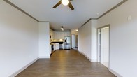 This single-floor unit showcases an open-concept layout that seamlessly connects the living area and kitchen, creating a spacious and functional environment. The flooring throughout consists of rich, dark hardwood-style planks that add warmth and a contemporary feel to the space.

The kitchen is outfitted with white cabinetry extending to the upper walls, providing ample storage. Stainless steel appliances, including a French door refrigerator, dishwasher, and gas range, offer a modern and cohesive aesthetic. Dark countertops contrast neatly with the white cabinetry, and track lighting above the kitchen ensures well-lit food preparation areas.

The open living area benefits from the same continuous flooring and features a ceiling fan with a globe light fixture, enhancing air circulation and ambient lighting. Crown molding runs along the upper perimeter of the walls, adding a refined architectural detail. The neutral, light grey walls throughout create a clean and versatile backdrop. A doorway visible at the far end suggests access to additional rooms, contributing to the overall functional flow of the space.