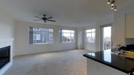 This single-floor space offers an open, airy layout designed to maximize comfort and functionality. The main living area is generously proportioned, with soft wall-to-wall carpeting underfoot and crisp white walls that create a clean, neutral backdrop throughout. A classic ceiling fan is centrally mounted overhead, providing both air circulation and a subtle decorative touch.

Natural light is a defining feature of this space. A series of large, multi-pane windows spans the living area, framing views of the surrounding residential buildings and a tree-lined backdrop beyond. The windows flood the interior with daylight, enhancing the sense of openness and creating a bright, welcoming atmosphere throughout the day.

Adjacent to the living area, a kitchen counter with a sleek dark granite or quartz surface is visible, hinting at a well-appointed kitchen that transitions seamlessly into the living space. This open-concept connection between the kitchen and living areas promotes an ideal flow for everyday living and entertaining.

The overall ambiance of the space is light, clean, and versatile, with neutral finishes that provide a blank canvas for personalization. The combination of abundant windows, open layout, and quality finishes makes this a comfortable and practical space.