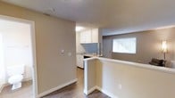 This single-floor studio unit offers an efficiently designed open-concept layout that combines the living and kitchen areas into one cohesive space. The kitchen is positioned toward the rear and features white cabinetry, a light blue tile backsplash, a stainless steel sink, and a dishwasher. A half-wall peninsula separates the kitchen from the main living area, providing a natural division while maintaining the open feel of the space. The flooring throughout is a wood-look laminate in a warm brown tone, adding visual warmth and continuity across the unit. Walls are finished in a neutral beige palette, complementing the white trim and baseboards. A textured ceiling adds subtle character overhead. A window near the entry door allows natural light to filter into the space, brightening the interior. The single bathroom completes the unit. Overall, the studio presents a clean, functional design well-suited for efficient everyday living.