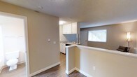 This single-floor studio unit offers an efficiently designed open-concept layout that combines the living and kitchen areas into one cohesive space. The kitchen is positioned toward the rear and features white cabinetry, a light blue tile backsplash, a stainless steel sink, and a dishwasher. A half-wall peninsula separates the kitchen from the main living area, providing a natural division while maintaining the open feel of the space. The flooring throughout is a wood-look laminate in a warm brown tone, adding visual warmth and continuity across the unit. Walls are finished in a neutral beige palette, complementing the white trim and baseboards. A textured ceiling adds subtle character overhead. A window near the entry door allows natural light to filter into the space, brightening the interior. The single bathroom completes the unit. Overall, the studio presents a clean, functional design well-suited for efficient everyday living.