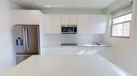 This single-floor space showcases a clean, contemporary design aesthetic centered around a beautifully appointed kitchen. The kitchen is defined by its minimalist approach, featuring floor-to-ceiling flat-panel white cabinetry that creates a seamless, uncluttered look along the full length of the wall. Stainless steel appliances, including a built-in microwave and a full-size refrigerator, provide a sharp contrast against the all-white palette, lending a professional and polished feel. A black ceramic cooktop is flush-mounted into the countertop, maintaining the streamlined appearance, while a stainless steel sink with a modern arc-neck faucet completes the functional workspace. The countertops throughout are a light-toned quartz, offering both durability and visual continuity. A generous kitchen island anchors the space, providing ample surface area for meal preparation or casual dining. The overall ambiance is bright and airy, enhanced by the crisp white surfaces that reflect natural light throughout. The space exudes a sense of order and simplicity, making it well-suited for those who appreciate modern, functional design with a refined, understated character.