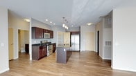 This single-floor space showcases a thoughtfully designed open-concept layout, with the kitchen serving as a central focal point. Rich, dark-stained wood cabinetry lines the kitchen walls, complemented by dark granite countertops that provide a striking contrast to the light hardwood flooring extending throughout the space. Stainless steel appliances, including a gas range with oven, microwave, and a side-by-side refrigerator, lend a modern, cohesive aesthetic to the kitchen. A center island with a built-in sink and matching dark countertop offers additional workspace and casual seating potential, while a trio of pendant lights overhead adds focused illumination and a design accent. Recessed lighting in the ceiling further enhances the brightness of the space. The open layout allows a clear sightline into the adjacent living areas, contributing to an airy, connected feel. Neutral gray walls and light flooring create a clean, contemporary backdrop. Multiple doorways visible from the kitchen suggest well-organized access to private rooms beyond, making the overall floor plan both practical and inviting.