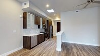 This single-floor space showcases a thoughtfully designed, modern kitchen as its centerpiece. The kitchen features rich, dark wood cabinetry paired with frosted glass upper cabinet inserts, creating a sophisticated contrast against the crisp white walls and light hardwood-style flooring that runs throughout. A classic white subway tile backsplash adds texture and a timeless touch to the cooking area.

The kitchen is equipped with a full suite of stainless steel appliances, including a gas range, built-in microwave, refrigerator, and dishwasher, catering to both everyday and gourmet cooking needs. A white quartz or similarly light-toned countertop provides ample workspace, complemented by a stainless steel sink with a modern gooseneck faucet.

Natural light is a standout feature, with a skylight positioned directly above the kitchen, flooding the space with daylight alongside recessed ceiling lighting. A partial white partition wall with a decorative column separates the kitchen from the adjacent open area, enhancing the sense of flow. A stylish arched dark wood door is visible in the background, adding an architectural accent to the otherwise clean, contemporary design.
