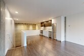 Located at 2358 Eastlake Ave E in Seattle's vibrant Eastlake neighborhood, this contemporary two-story unit offers a thoughtfully designed layout spread across two levels. The upper floor serves as the main living and kitchen area, showcasing an open-plan design with warm hardwood-style flooring throughout. The kitchen is outfitted with stainless steel appliances—including a range, microwave, refrigerator, and dishwasher—complemented by a two-tone cabinet scheme of white lower cabinets and rich dark wood uppers, plus a clean white countertop. A track-lit accent wall with a textured metallic finish adds an industrial-chic character, while exposed concrete columns and a large street-facing window flood the space with natural light. A central peninsula defines the kitchen boundary and provides additional workspace. The lower floor houses the living room, which features exposed concrete walls that reinforce the space's urban aesthetic, along with the full bathroom, a spacious walk-in closet with wire shelving, an in-unit stacked washer/dryer, and a hallway connecting the amenities. The bathroom is finished in crisp white with a tub/shower combo and a wide vanity. A staircase connects both levels seamlessly, completing this well-appointed urban residence.