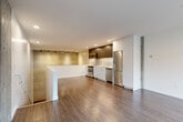Located at 2358 Eastlake Ave E in Seattle's vibrant Eastlake neighborhood, this contemporary two-story unit offers a thoughtfully designed layout spread across two levels. The upper floor serves as the main living and kitchen area, showcasing an open-plan design with warm hardwood-style flooring throughout. The kitchen is outfitted with stainless steel appliances—including a range, microwave, refrigerator, and dishwasher—complemented by a two-tone cabinet scheme of white lower cabinets and rich dark wood uppers, plus a clean white countertop. A track-lit accent wall with a textured metallic finish adds an industrial-chic character, while exposed concrete columns and a large street-facing window flood the space with natural light. A central peninsula defines the kitchen boundary and provides additional workspace. The lower floor houses the living room, which features exposed concrete walls that reinforce the space's urban aesthetic, along with the full bathroom, a spacious walk-in closet with wire shelving, an in-unit stacked washer/dryer, and a hallway connecting the amenities. The bathroom is finished in crisp white with a tub/shower combo and a wide vanity. A staircase connects both levels seamlessly, completing this well-appointed urban residence.