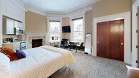 Located at 888 Delaware Ave in Buffalo, NY, this one-bedroom, one-bathroom suite blends historic charm with modern functionality. The spacious main room is defined by elegant architectural details, including ornate crown molding, a decorative bay-style window alcove, and a striking marble fireplace that serves as a focal centerpiece. Warm beige walls complement the neutral carpeted flooring, creating a cohesive and inviting atmosphere throughout. Large double-hung windows allow abundant natural light to fill the room while offering views of the surrounding neighborhood's classic brick architecture. The main room is thoughtfully arranged to accommodate a king-sized bed, a dedicated workspace with a wall-mounted television, and a compact kitchenette equipped with upper cabinetry, a microwave, and a small range — providing essential conveniences within the space. A full-length mirror and leather accent chair add practical and aesthetic touches. The bathroom features marble-look tile flooring, a vanity with ample cabinet storage, a well-lit mirror, and a glass-enclosed shower, maintaining a clean and polished aesthetic. A rich-toned solid wood door anchors the entryway, reinforcing the suite's historic character. This space offers a well-preserved blend of period architectural details and contemporary amenities.