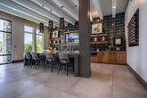 Located at 1980 Icon Way in Knoxville, Tennessee, this single-story amenity facility offers a thoughtfully designed space that blends contemporary style with practical functionality. The grand entrance lobby sets the tone with soaring ceilings, large-format tile flooring, an eye-catching geometric chandelier, and a striking warm wood accent wall. Abundant natural light streams through floor-to-ceiling windows and glass entry doors, creating a bright and welcoming atmosphere. A well-appointed coffee and refreshment bar features warm wood cabinetry, a bold geometric tile backsplash, open shelving, and a mounted TV — ideal for casual gatherings. The lounge area offers a sophisticated retreat with deep blue sectional seating, an integrated fireplace set within a sleek dark surround, exposed beam ceilings, and direct views of the outdoor pool area. The expansive fitness center is a standout, boasting soaring ceilings, walls of oversized windows with outdoor views, hardwood-style flooring, and a comprehensive array of equipment including treadmills, ellipticals, rowing machines, cable systems, free weights, and a heavy bag. A built-in shelving unit stocks towels and accessories for convenience. Water bottle filling stations with textured tile surrounds complete the thoughtful amenities of this well-equipped facility.