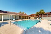 Located at 10530 Florida Blvd in Baton Rouge, Louisiana, this outdoor pool facility offers a well-appointed recreational space designed for relaxation and leisure. The centerpiece is a generously sized swimming pool with crystal-clear blue water, accessible via dual stainless steel entry rails and featuring wide entry steps at the shallow end, making it welcoming for all swimmers.

The pool deck is finished in a light-toned, textured surface that extends around the full perimeter of the pool, providing ample space for sunbathing and socializing. Multiple turquoise lounge chairs are positioned throughout the deck, offering comfortable spots to unwind under the open Louisiana sky.

Bordering one side of the pool is a covered patio structure with a classic pitched roof supported by white columns. This shaded area provides a cool retreat from the sun and appears to connect to an interior facility space, offering additional amenities nearby. The surrounding enclosure features a sturdy brick wall with decorative lattice inserts, lending the area a sense of privacy and a distinct architectural character.

Mature palm trees and tall pines frame the space, enhancing the tropical, resort-like atmosphere. The property enjoys abundant natural light under clear skies, further elevating the outdoor experience. The overall setting is clean, well-maintained, and thoughtfully designed for communal outdoor enjoyment.