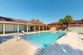 Located at 10530 Florida Blvd in Baton Rouge, Louisiana, this outdoor pool facility offers a well-appointed recreational space designed for relaxation and leisure. The centerpiece is a generously sized swimming pool with crystal-clear blue water, accessible via dual stainless steel entry rails and featuring wide entry steps at the shallow end, making it welcoming for all swimmers.

The pool deck is finished in a light-toned, textured surface that extends around the full perimeter of the pool, providing ample space for sunbathing and socializing. Multiple turquoise lounge chairs are positioned throughout the deck, offering comfortable spots to unwind under the open Louisiana sky.

Bordering one side of the pool is a covered patio structure with a classic pitched roof supported by white columns. This shaded area provides a cool retreat from the sun and appears to connect to an interior facility space, offering additional amenities nearby. The surrounding enclosure features a sturdy brick wall with decorative lattice inserts, lending the area a sense of privacy and a distinct architectural character.

Mature palm trees and tall pines frame the space, enhancing the tropical, resort-like atmosphere. The property enjoys abundant natural light under clear skies, further elevating the outdoor experience. The overall setting is clean, well-maintained, and thoughtfully designed for communal outdoor enjoyment.