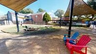 Located at 1700 Seaspray Ct in Houston's 77008 zip code, this thoughtfully designed outdoor dog park offers a welcoming and functional space for pets and their owners. The single-level grounds are enclosed by a sturdy horizontal-slat wood and metal fence, providing a secure perimeter that ensures safety while maintaining an open, airy feel.

Multiple shade sails are installed throughout the space, suspended from sleek black steel posts, offering generous relief from the Texas sun across several distinct activity zones. Colorful Adirondack chairs in blue and red are positioned beneath the canopies, providing comfortable seating for visitors to relax while their pets roam freely.

The park features a variety of ground surfaces, including natural gravel and packed earth, ideal for pet activity. Lush tropical plantings — including large bird's nest ferns and ornamental shrubs — line the perimeter, creating a vibrant green border that softens the fencing and adds natural beauty. Mature oak and deciduous trees frame the space overhead, contributing additional shade and a park-like atmosphere.

A dedicated entry gate with posted signage provides controlled access, and log obstacles and natural rock features are scattered across one section of the grounds, offering enrichment elements for dogs. The surrounding brick residential buildings are visible beyond the fence, situating the park within a larger community environment.
