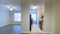 This single-story residence showcases an open-concept design that connects the kitchen and living areas in a seamless, functional layout. The kitchen is equipped with black appliances, including a gas range and refrigerator, complemented by warm wood-toned cabinetry and an under-cabinet range hood. A flush-mount fluorescent light fixture illuminates the kitchen workspace, while the flooring transitions from light-toned vinyl or laminate in the kitchen to hardwood-style flooring in the living area. Structural columns with crown molding detailing define the separation between spaces without fully enclosing them, maintaining an open feel throughout. The living area benefits from multiple windows fitted with horizontal blinds, allowing ample natural light to fill the space. Crown molding runs consistently along the ceilings, adding a refined architectural touch, while neutral warm-gray walls provide a versatile backdrop. The textured ceilings and clean white baseboards complete the interior aesthetic. The overall ambiance is bright, practical, and well-suited for everyday living, with a cohesive design that balances function and comfort across the connected spaces.