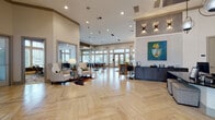 This single-floor clubhouse is a thoughtfully designed community facility that seamlessly blends social and functional spaces. The expansive lobby greets visitors with striking herringbone-patterned light wood flooring, soaring ceilings, and recessed lighting throughout. A prominent reception desk anchors one side of the lobby, complemented by tasteful seating areas and bold contemporary artwork, including a vivid portrait painting that serves as a focal point on a textured accent wall.

The lounge area features a warm, wood-paneled accent wall with a mounted flat-screen television, a circular chandelier, plush sectional seating, and an eclectic mix of accent chairs atop patterned carpet. Large windows flood the space with natural light and offer views of the surrounding landscape.

A dedicated conference room provides a polished environment for meetings, outfitted with a substantial wood table, upholstered chairs, tall windows, and refined wall art beneath a dark tray ceiling. Adjacent private offices with glass partitions offer flexibility for leasing or administrative use.

The game room extends the social atmosphere with a large bar-height island, shuffleboard table, foosball, and floor-to-ceiling windows. A well-appointed kitchenette with white quartz countertops, stainless steel appliances, and herringbone tile flooring rounds out this versatile and welcoming space.