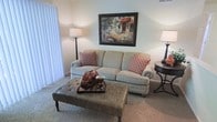 This single-floor space features a thoughtfully designed living room that balances comfort and style. The room is anchored by a plush, light-toned sofa accented with decorative patterned throw pillows in warm earth tones, complementing the neutral color palette of the walls and soft carpeted flooring. A tufted leather ottoman serves as a functional centerpiece, paired with a decorative tray displaying artistic accents. Ambient lighting is provided by two floor and table lamps positioned on either side of the seating area, creating a warm and inviting atmosphere. A framed landscape painting mounted above the sofa adds a classic artistic touch to the space. Vertical blinds along the window allow for flexible light control while providing privacy. A small round side table with elegant curved legs completes the seating arrangement. The overall ambiance is warm, cozy, and traditionally styled, making the living area feel welcoming and well-appointed.