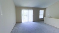 This single-floor space features a spacious, open living area finished with wall-to-wall carpeting in a neutral gray tone, contributing to a clean and understated aesthetic. The room is characterized by its simplicity and functionality, with smooth, light-beige walls that create a warm, airy atmosphere. A large sliding glass door, dressed with vertical blinds, serves as the focal point of the main living area, allowing abundant natural light to fill the space while providing access to an outdoor area. An additional window on the adjacent wall further enhances the brightness of the room, ensuring consistent natural illumination throughout the day. The layout is open and unobstructed, offering flexibility for a variety of furniture arrangements. A partial wall or countertop is visible toward one side of the room, suggesting a transition into a separate kitchen or dining area. The overall design is straightforward and practical, with neutral finishes throughout that provide a versatile backdrop suitable for a range of interior styles and personal preferences.
