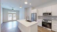 Located in Meadows Place, TX, this thoughtfully designed one-bedroom, one-bathroom residence offers a clean, modern aesthetic across a single floor. The open-concept layout seamlessly connects the kitchen and living area, maximizing the sense of space and flow. The kitchen is a standout feature, showcasing crisp white shaker cabinetry, a light gray tile backsplash, stainless steel appliances including a French door refrigerator, built-in microwave, and gas range, as well as a generous center island with a white quartz countertop, an undermount stainless steel sink, and a built-in dishwasher. Pendant lighting and recessed fixtures provide ample illumination, while warm hardwood-style flooring anchors the main living areas. A ceiling fan and a windowed entry door allow natural light to filter into the living space. The bedroom is carpeted for comfort and features panel-style doors with direct access to both the bathroom and a dedicated closet. The bathroom is well-appointed with wood-look flooring, a white vanity with quartz countertop, an LED-backlit mirror, and convenient access to the walk-in closet with built-in shelving. Neutral tones, clean lines, and modern hardware unify the space with a polished, contemporary character throughout.