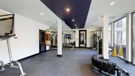 This two-floor fitness facility is designed to accommodate a comprehensive range of training disciplines. The main workout floor features a striking design with crisp white walls contrasted by deep navy accent panels on the ceiling, creating a bold and energizing atmosphere. Recessed lighting illuminates the space evenly throughout, while dark rubberized flooring provides both durability and safety underfoot. The facility is well-equipped for diverse training styles. A leather heavy punching bag hangs from the ceiling near the entrance, catering to combat and cardio training. Strength training is supported by a multi-station cable machine, a weight rack stocked with an array of dumbbells and plates, and additional resistance equipment. A large tractor tire on the floor signals a dedicated functional fitness and conditioning zone. Mirrored walls extend the visual depth of the space and are a practical addition for form-checking during workouts. A mounted flat-screen television keeps members entertained or informed during sessions. Structural columns are neatly integrated into the layout without disrupting the flow of the workout areas. The overall ambiance is clean, professional, and purposefully laid out to support both individual training and small group sessions across its two levels.
